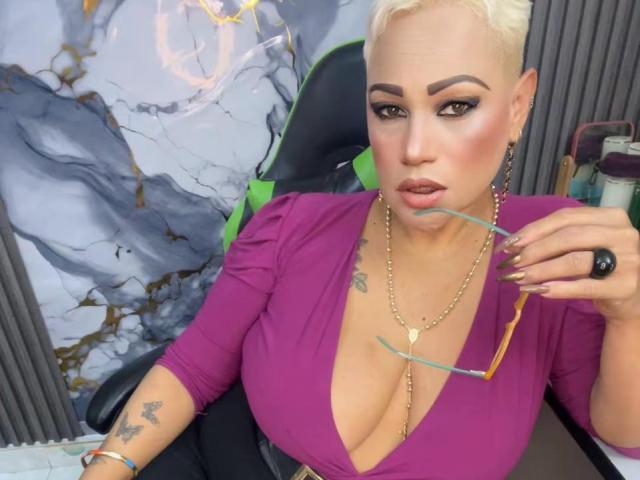 GianellaCox, avec une coupe de cheveux courte blonde, portant un haut violet à col en V plongeant, montrant de grandes seins, tenant des lunettes dans la bouche, un collier en or, une chaise noire, des accents verts, des tatouages sur le bras..