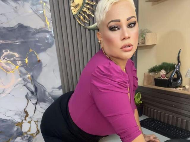 Gianella Cox, con i capelli biondi corti, indossa una camicia rosa, pantaloni neri e orecchini, è appoggiata a una scrivania, mostrando il sedere, e guarda oltre la spalla con il trucco..