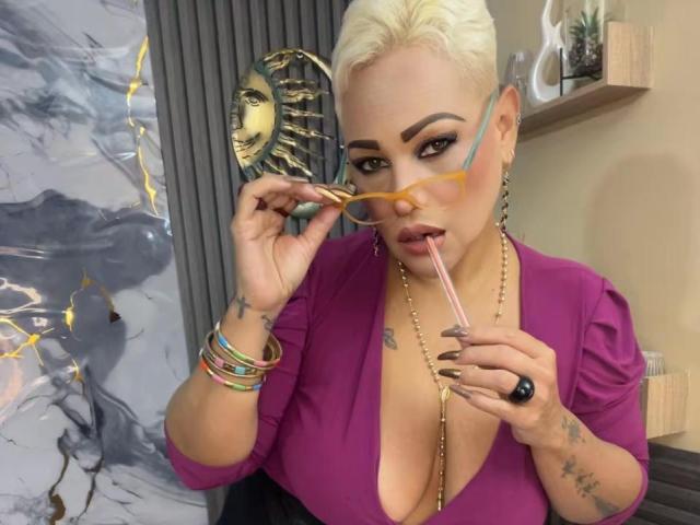 Gianella Cox, con i capelli biondo platino, indossa una maglietta viola, tiene degli occhiali rosa vicino alle labbra, mostrando il suo seno prosperoso, tatuaggi sulle mani e braccialetti colorati..