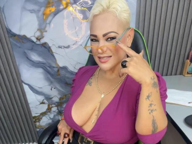 GianellaCox with short blonde hair, big tits, wearing a purple top, gold necklace, and glasses, pointing at them with a smirk, tattoos on her right hand.