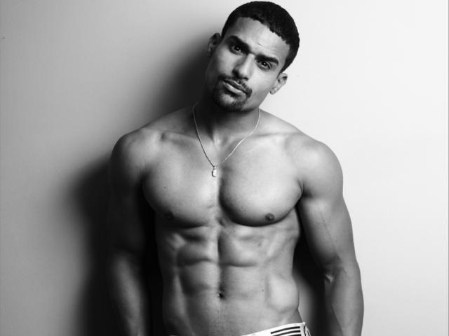 AdonisDios est un homme noir musclé, avec une courte coupe de cheveux, portant un collier et des sous-vêtements blancs, montrant des abdominaux bien définis et une expression sérieuse sur une photo en noir et blanc..