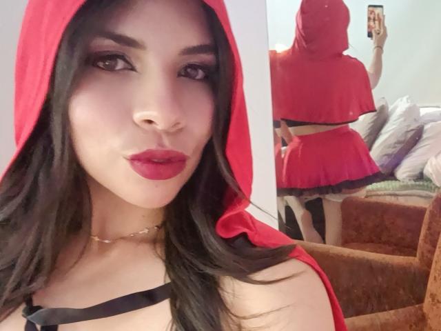 VioletaStone con capucha roja, correa negra y falda roja, labios fruncidos, el reflejo en el espejo muestra su trasero, cabello negro largo, lápiz labial rojo, collar dorado, sillón marrón, dormitorio..