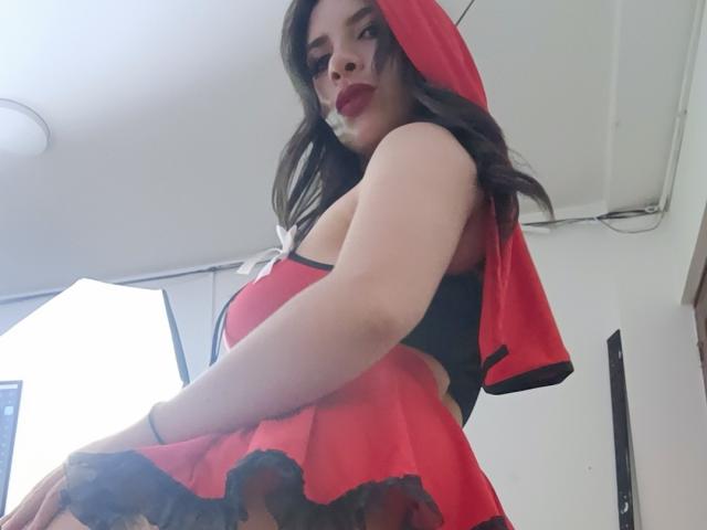 VioletaStone com capuz vermelho, renda preta e saia vermelha, mostrando os seios e o bumbum, maquiagem escura e batom vermelho, olhando para baixo, em um plano de ângulo baixo..