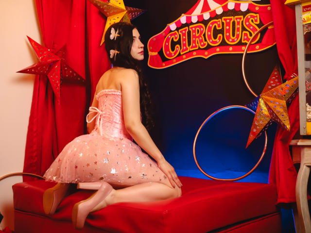 Jade Harpper, con un vestido rosa, brillante y sin tirantes, arrodillada sobre un cojín rojo con las nalgas levantadas, cabello negro largo, un tocado con forma de estrella, tacones rosas, cortinas rojas y un fondo con temática de circo..