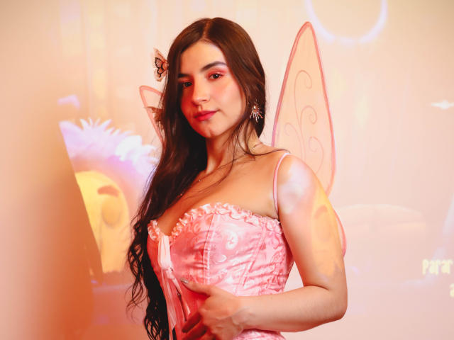 Jade Harpper, con i suoi lunghi capelli castani, le ali da fata rosa e un corpetto rosa aderente, che mette in mostra il suo seno grande e formoso, con un sorriso sexy e sicuro e un piccolo piercing al naso..