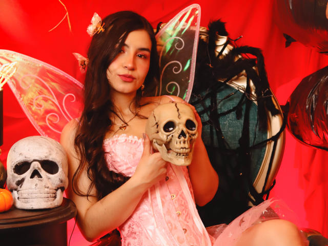 Jade Harpper, con i suoi lunghi capelli scuri, indossa un abito di pizzo rosa e delle ali da fata, tiene in mano un teschio, è seduta a un tavolo con un altro teschio e una borsa di pelle nera..