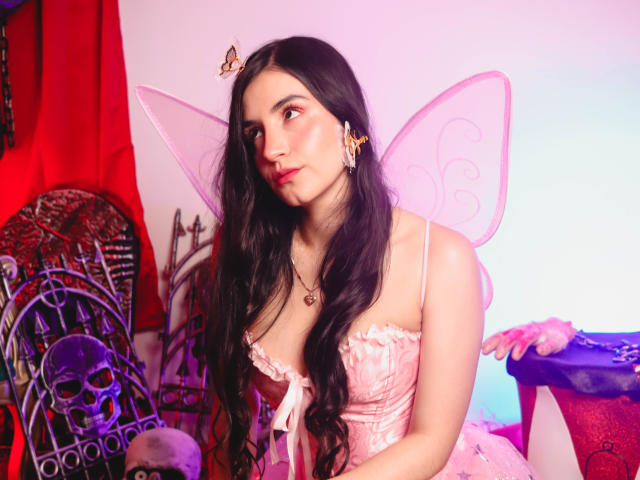 JadeHarpper with long black hair, pink fairy wings, and a pink corset, showing her tits, wearing a butterfly hairclip, sitting near a skull chair, looking sexy.