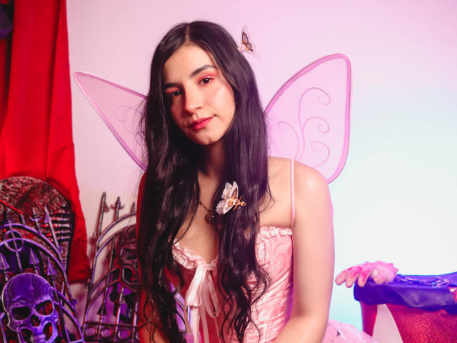 Jade Harpper, con lunghi capelli neri, ali da fata rosa e una forcina a forma di farfalla, indossa una camicetta di pizzo rosa, sorride, ha un viso carino e piccoli seni. Una farfalla sulla sua testa..