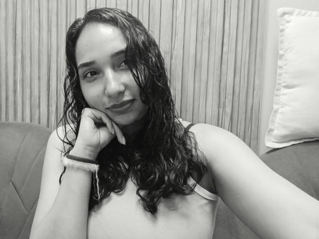 Black-and-white selfie of EleanorLamberth with wet, curly hair, resting her chin on her hand, wearing a bra, looking at the camera, smiling subtly, wrist with a fuzzy bracelet.
