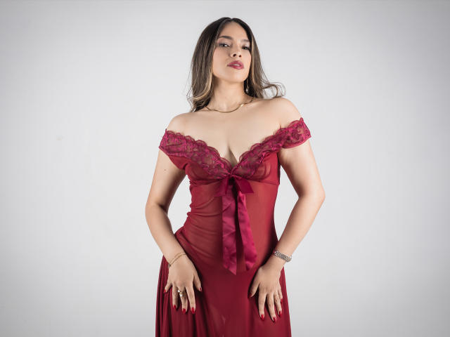 VivienneLaurenttte con un vestido rojo de encaje con un lazo, de corte asimétrico, de pie con las manos en las caderas, mostrando sus senos, cabello castaño y lápiz labial rojo..