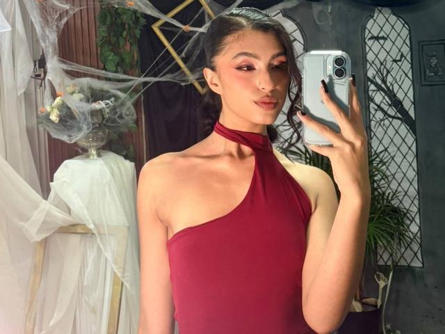 LilithDesire, avec ses cheveux noirs attachés en queue de cheval, portant une robe rouge à encolure dégagée, tenant un téléphone, prenant un selfie devant un miroir, montrant un visage mince et mignon et de petites seins..