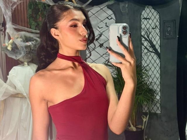 LilithDesire, con i capelli scuri raccolti in una coda di cavallo, indossa un vestito rosso a halter, tiene in mano un telefono e sta scattando un selfie allo specchio, mostrando un busto slanciato, carino e sexy..