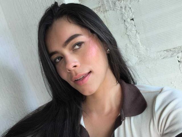 AlanaBlake with long black hair, brown eyes, pink blush on cheeks, small nose, slight smile, wearing white shirt with brown collar, ear piercing, against textured white wall.