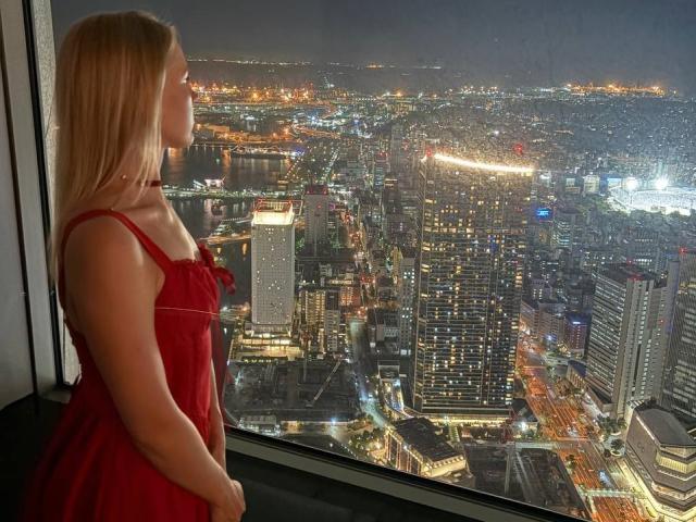 DaneteHeingartner with blonde hair, red dress, standing by window, looking down at city at night, visible ass, city lights, tall buildings, river, reflections, high-rise, urban skyline.
