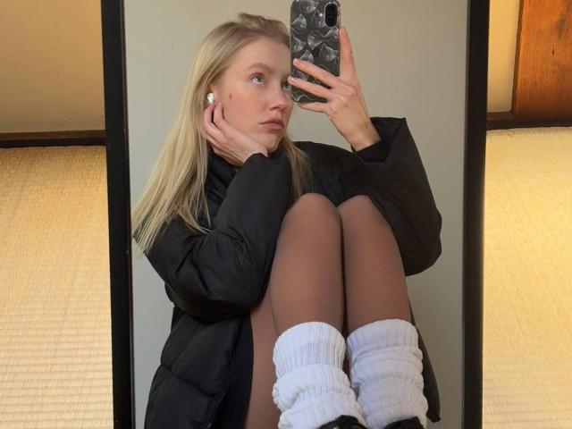 DaneteHeingartner with blonde hair, wearing a black puffy coat, white socks, and black shoes, sitting with knees up, taking a mirror selfie, showing brown panty legs.