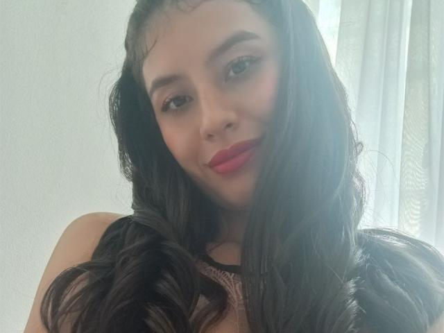 MadissonFontaine has long, wavy black hair, pink lipstick, and is smiling slightly at the camera. Her brown eyes are looking forward. She's wearing a black lace top.