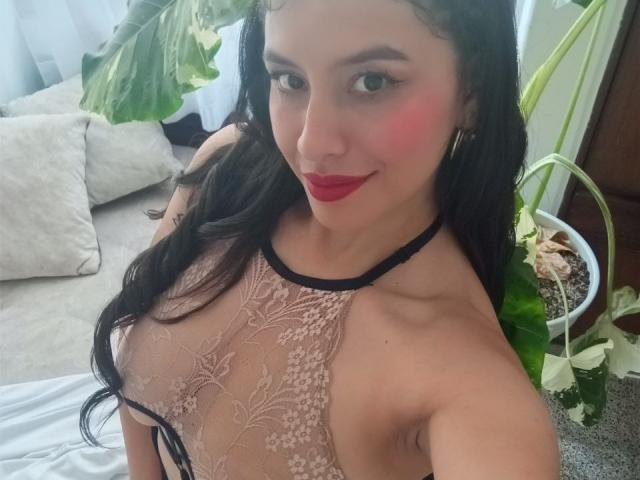 MadissonFontaine with long black hair, red lips, and pink cheek, wearing a sheer lace top that reveals her large tits, standing near a potted plant, smiling at the camera.