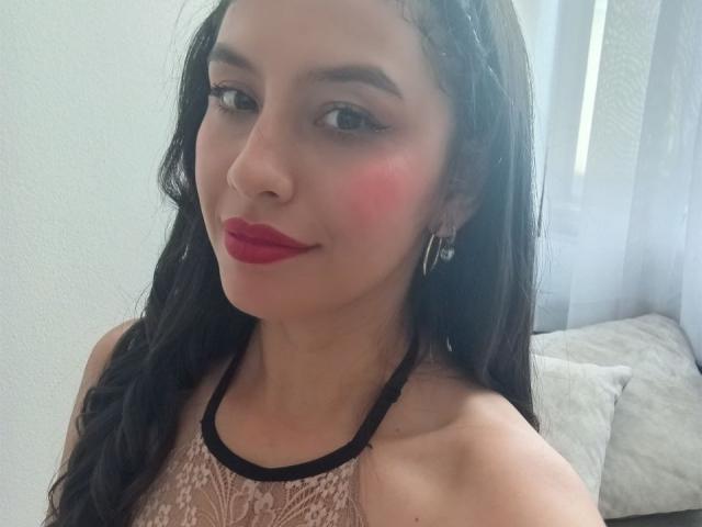 MadissonFontaine has dark hair, red lipstick, and pink blush, wearing a lace halter top with black straps, and small hoop earrings, smiling at the camera.