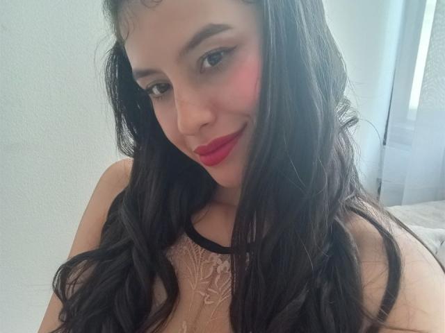 MadissonFontaine has long black hair, red lipstick, and is wearing a sheer lace top, smiling at the camera with a slight blush on her cheeks. Her tits are partially visible.