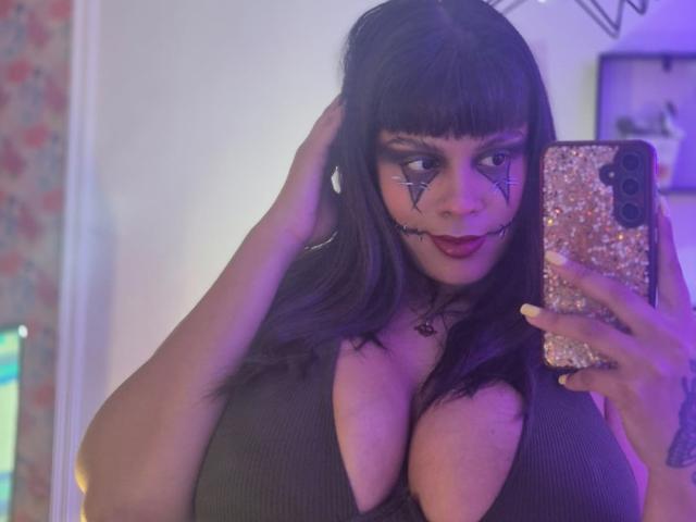 TeylorJenkins with dark hair, wearing a deep cleavage gray top, face painted with black triangle and line designs, holding a glittery phone, purple-lit room, big tits.