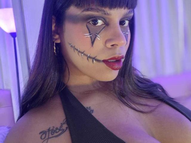 TeylorJenkins has dark hair, red lipstick, and a black eye makeup with star and stitch tattoos on her face. She's wearing a black halter top, showing her tits.