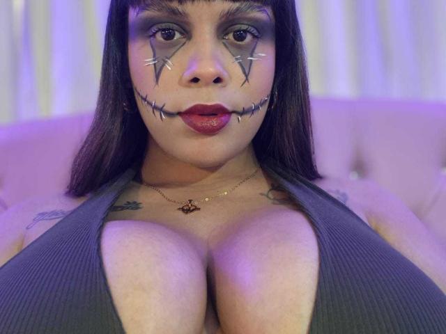 TeylorJenkins has dark makeup with zombie eyes, red lipstick, and a deep cleavage in a gray top. Her large tits are prominent, and she's wearing a necklace.