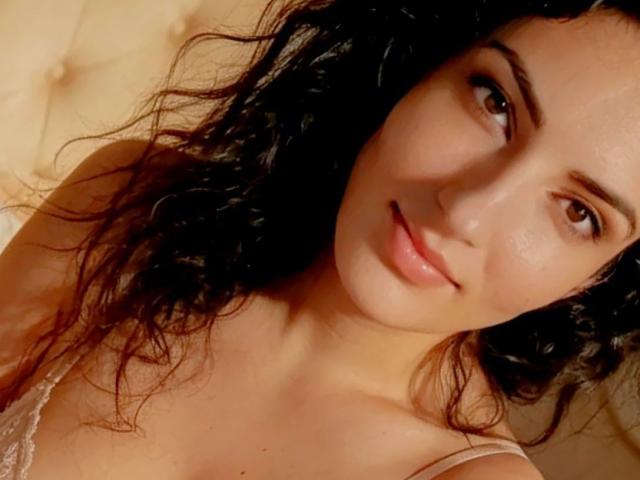 AddictedToUTS with curly black hair, light skin, brown eyes, and a subtle smile, wearing a lace top, looking up at the camera. Her tits are slightly visible.