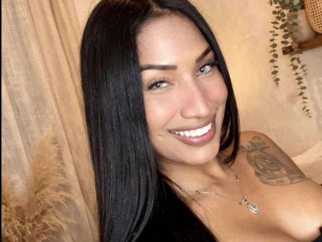 HannaMarceau with long black hair, smiling, showing white teeth, tan skin, green eyes, and a rose tattoo on her right shoulder, wearing a necklace, in a warm-lit photo.
