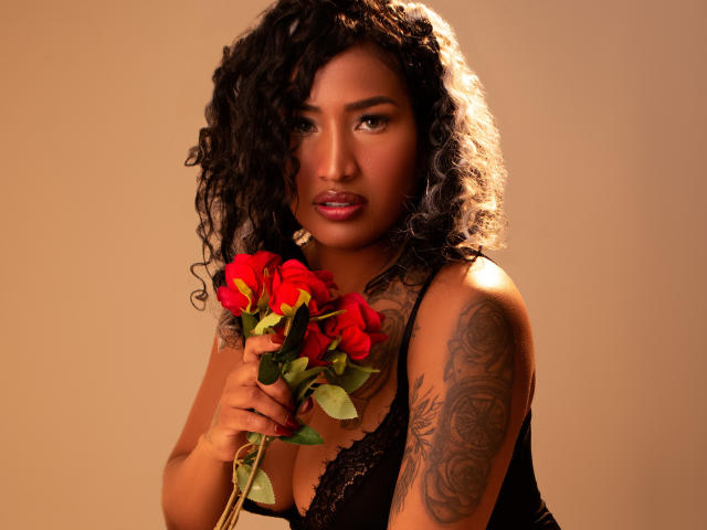 HannaMarceau with curly black hair, holding red roses, wearing black lace lingerie, showing a tattoo on her right arm, brown skin, and looking seductively at the camera.