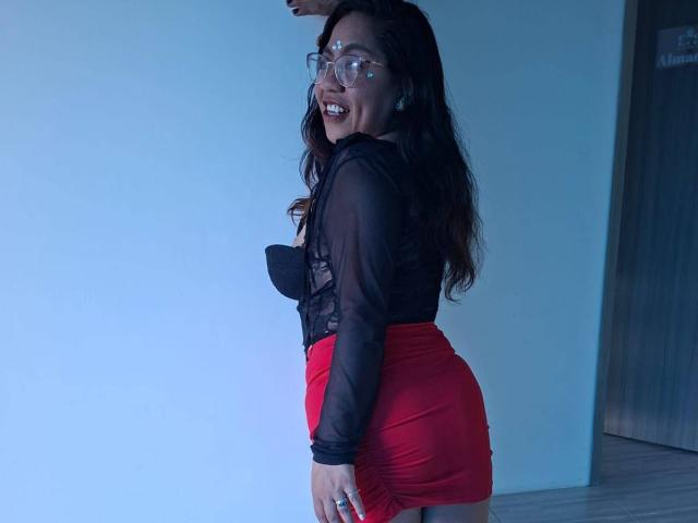 SilvanaLane with long dark hair, glasses, black sheer top showing her tits, and red skirt, smiling, facing left, holding a wall. Blue light.