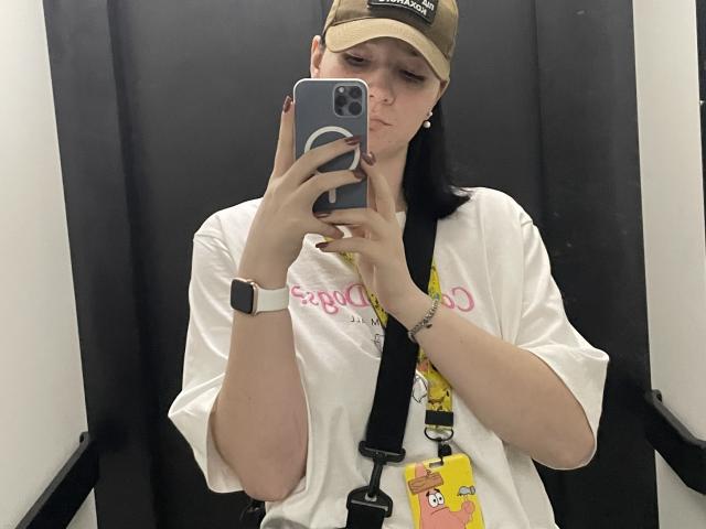 Ellaria with pale skin, black hair in a ponytail, wearing a beige cap, white shirt, and yellow lanyard, taking a mirror selfie with a serious expression, holding a smartphone.