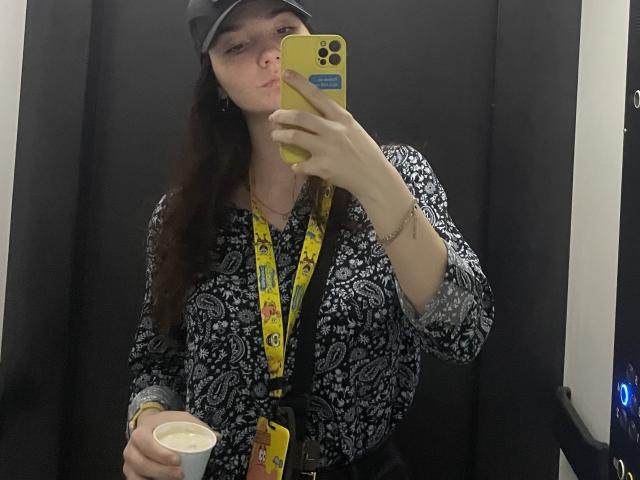 Ellaria with long brown hair, wearing a black cap, patterned blouse, yellow lanyard, holding a cup, taking a mirror selfie with a yellow phone, pouting her lips.