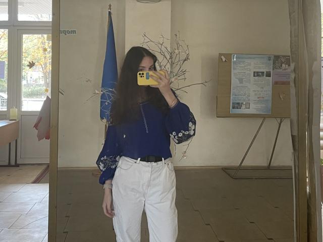 Ellaria with long brown hair, wearing a blue blouse with white floral sleeves and white high-waisted pants, taking a mirror selfie with a yellow phone, covering her face.