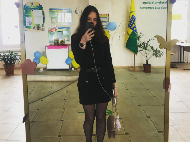 Ellaria with long brown hair, wearing a black dress, belt, and black patterned tights, taking a mirror selfie, holding a white bag, wearing a black watch.
