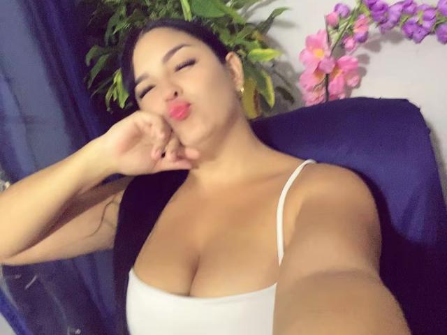 TalianaSquirt with pouty lips, hand near face, black hair, white tank top, showing big tits, cute, relaxed, eyes closed, selfie, pink flowers in background.