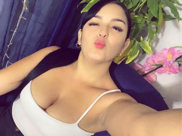 TalianaSquirt with pouty lips, black hair, hoop earrings, white tank top showing big boobs, black vest, taking selfie, green leafy plant in background, pink flowers on right.