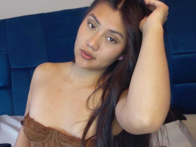 Catalehia69 with long brown hair, light skin, and brown eyes, wearing a lace strapless top, hand in hair, subtle nose ring, and soft expression, sitting on a blue bed.