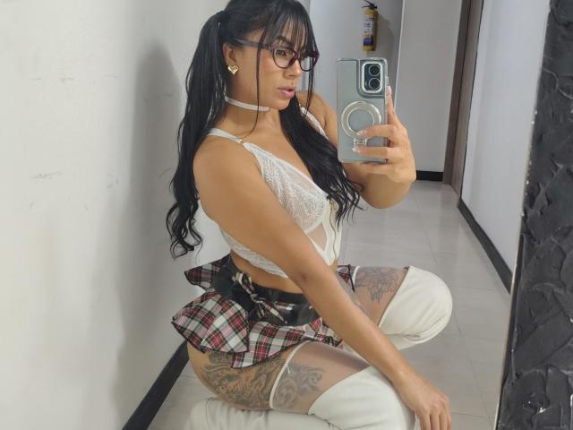 Amberv with long black hair, glasses, white lace bra, plaid skirt, white thigh-highs, and tattoos, kneeling, taking a mirror selfie, showing her ass and tattoos.