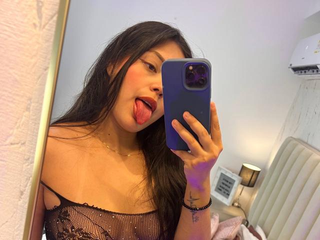 Violetta Rouse, con cabello negro largo, sacando la lengua, vistiendo una blusa negra de malla, sosteniendo un teléfono azul, tomando una selfie en el espejo, mostrando sus pequeños senos y su cuello delgado..