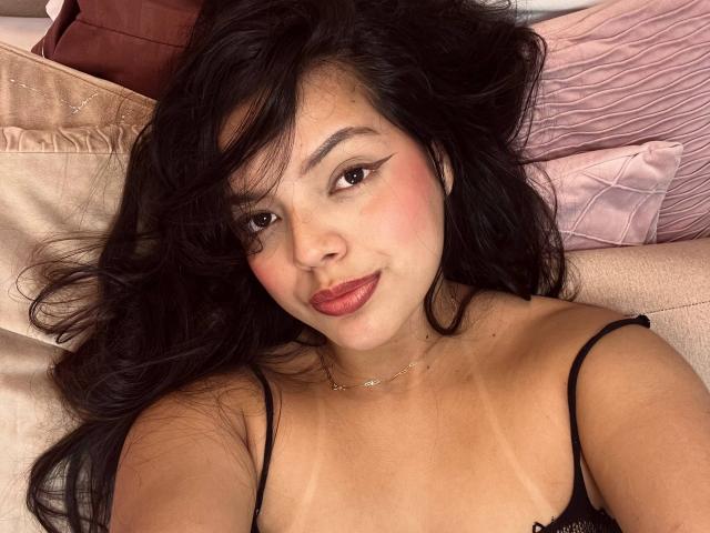 Violetta Rouse hat langes, schwarzes Haar, rosigen Lippen und roten Lippenstift. Sie liegt auf einem Bett mit beigen Kissen und trägt ein schwarzes Top mit Trägern, das ihre Brüste und Sonnenlinien zeigt..