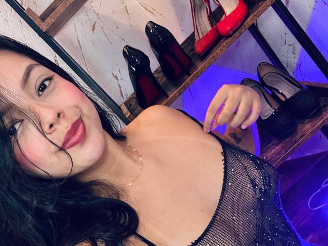 Violetta Rouse, con cabello oscuro, lápiz labial rojo y un vestido de malla negra, mostrando sus senos, sosteniendo zapatos de tacón negros, zapatos de tacón rojos en el fondo y luz púrpura..