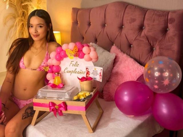 KittyMiller in pink lingerie, small tits, sitting on bed with pink headboard, next to gift box, pink balloons, and fairy lights, with a sign that says "Feliz Cumpleaños".