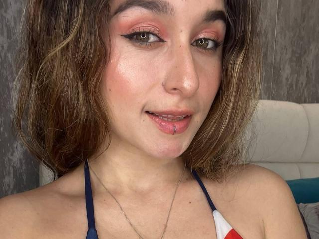 MilaGonz tiene un piercing en la nariz, un piercing en el labio y lleva un top azul con tirantes y un estampado rojo y blanco. Su maquillaje incluye sombra de ojos rosa y delineador de ojos negro..