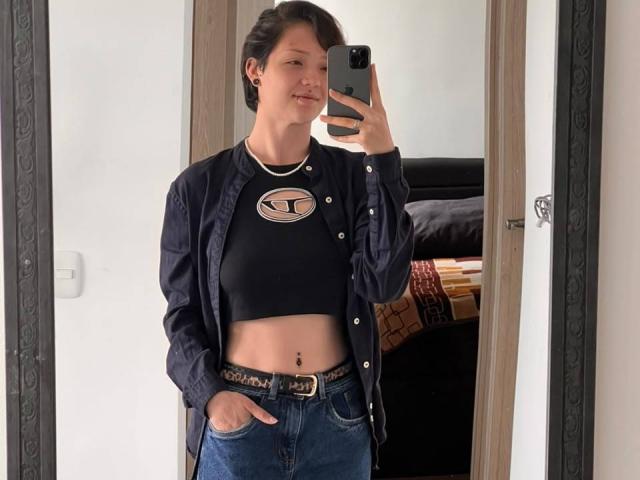 LissyLuxe with short black hair, wearing a black crop top, button-up shirt, and blue jeans, taking a mirror selfie, showing a belly button piercing and a belt with a buckle.