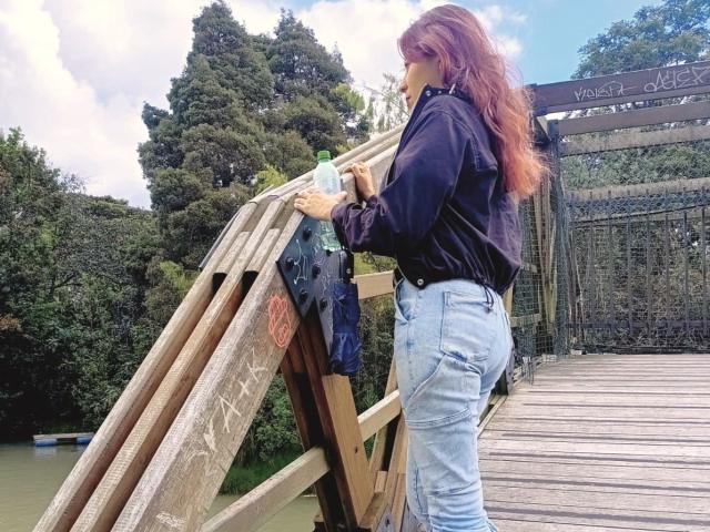 LunaTomson met lang rood haar, een zwart shirt en blauwe jeans, die een fles vasthoudt, staat op een houten brug, kijkt eromhoog en toont haar achterwerk..