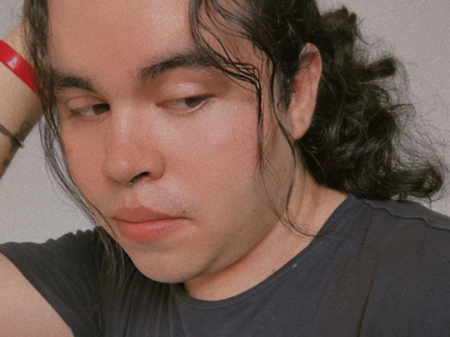 AngelRousse has a puffy ass face, slightly dirty skin, and a red wristband on his right arm, with messy black hair and a serious expression. Wearing a black shirt.