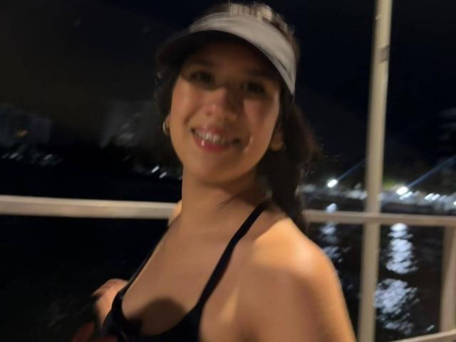 HannaThorne has dark hair, a black visor, and a black sports bra, showing off her small tits and smiling with her teeth slightly visible. Nighttime, blurry image.