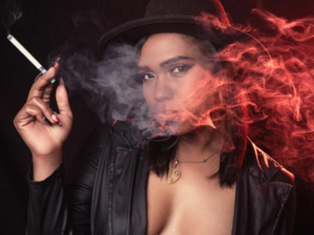 AntonellaVargas with smoky red and gray smoke, holding a cigarette, wearing a black hat and leather jacket, exposing her tits, black nails, and a gold necklace.
