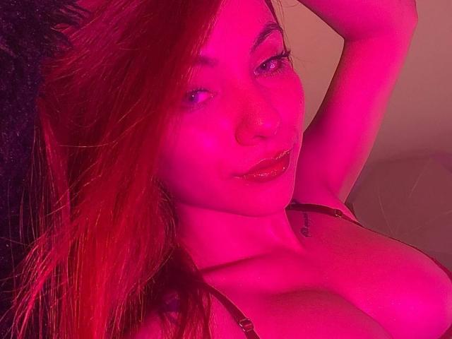 ValeryHarllow with pink-red lighting, raising arm, showing large boobs in a strapless top, with long red hair, glossy lips, and a small tattoo on the right side of neck.