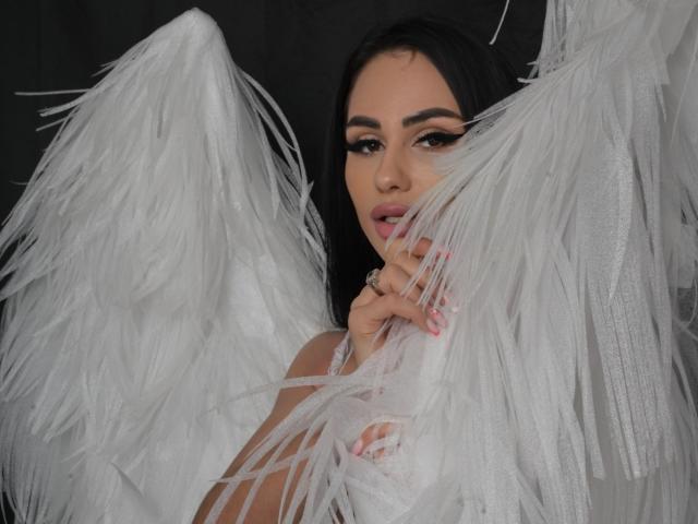 CharlotteAria with white feather wings, black hair, and pink lipstick, covering her tits with her hand, looking seductively at the camera. Her eyes are half-open and lips parted.
